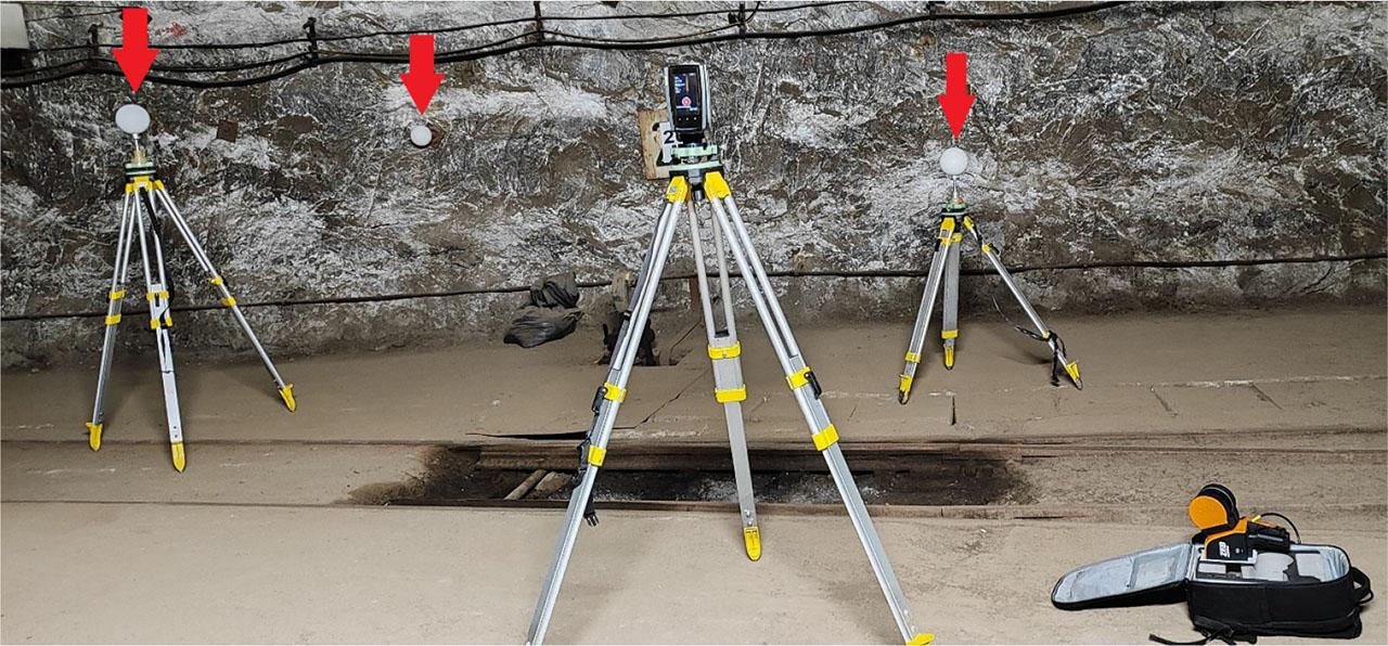Measurement site with a laser scanner in a mining excavation. A stationary scanner is visible in the central part of the photo. In the lower right corner, there is a mobile scanner intended to perform the measurement. In the background, three measuring spheres are marked with red arrows – two spheres placed on tripods under the geodetic network points and one additional sphere attached to the side of the excavation.